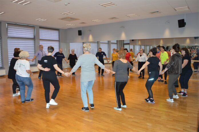 Tance Greckie_4 Grupa osób w sali tanecznej JDK stojąc w kółku trzyma się za ręce i uczy się kroków tańca greckiego.