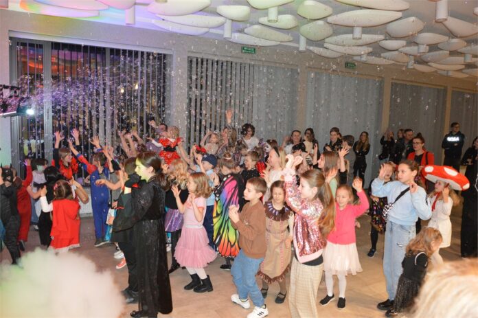 Obraz zawiera: Dzieci w kostiumach balowych wraz z rodzicami bawią się wraz z Lady Famą łapiąc bańki mydlane.