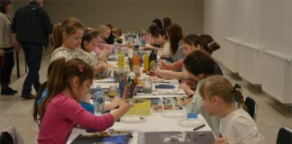 Malarskie podróże w JDK Obraz zawiera: Dzieci siedzą po obu stronach długiego stołu i malują obrazy, różnego rodzaju farbami i kredkami.