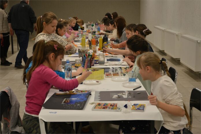 Malarskei podroze w JDK_1 Obraz zawiera: Dzieci siedzą po obu stronach długiego stołu i malują obrazy, różnego rodzaju farbami i kredkami.