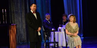 Obraz zawiera: Na pierwszym planie z lewej strony stoi mężczyzna w eleganckim płaszczu i w czerwonej muszce. W dłoniach trzyma rękawiczki. Z prawej strony na krześle siedzi kobieta i spogląda w jego stronę. W tle, za nimi przy kawiarnianym stoliku siedzi para.