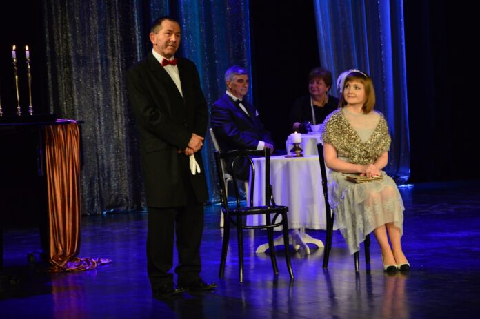 Powrocmy la za dawnych lat_11 Obraz zawiera: Na pierwszym planie z lewej strony stoi mężczyzna w eleganckim płaszczu i w czerwonej muszce. W dłoniach trzyma rękawiczki. Z prawej strony na krześle siedzi kobieta i spogląda w jego stronę. W tle, za nimi przy kawiarnianym stoliku siedzi para.