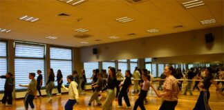 Feryjny hip hop Obraz zawiera: Grupa młodych dziewczyn wraz z instruktorką w sali tanecznej uczą się układu kroków w Hip Hopie, wychylając prawą nogę w bok.