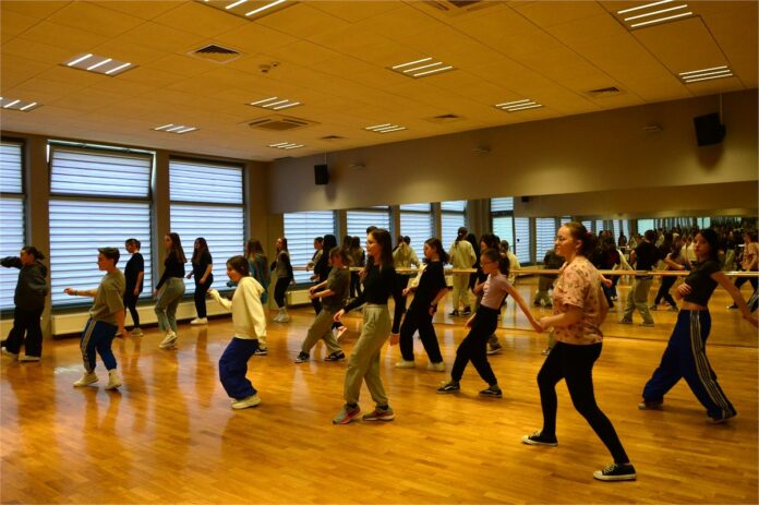 Warsztaty hip hop 2024_3 Obraz zawiera: Grupa młodych dziewczyn wraz z instruktorką w sali tanecznej uczą się układu kroków w Hip Hopie, wychylając prawą nogę w bok.