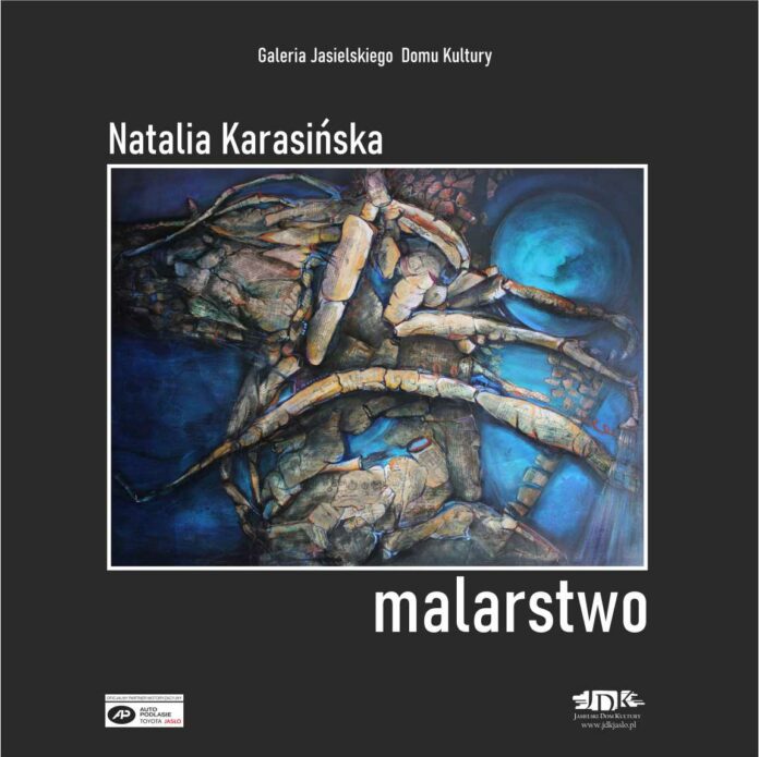 Obraz kwadratowy, czarne tło, z białymi napisami informującymi o nazwisku autora i miejscu wystawy. na środku obraz dziwnego tworu z niebieskim tłem. W lewym dolnym rogu logo tojoty, w prawym dolnym rogu logo jasielskiego domu kultury