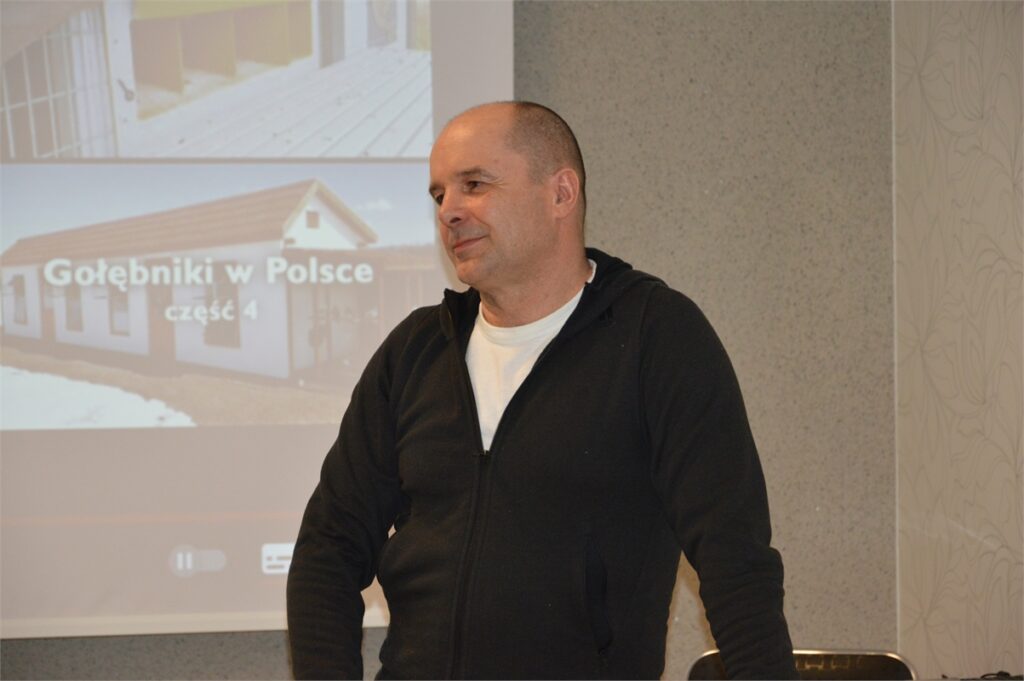 Obraz zawiera: Krótko obcięty mężczyzna w średnim wieku ubrany w ciemną bluzę polarową stoi na tle ekranu projekcyjnego, na którym pokazane są gołębniki w Polsce.