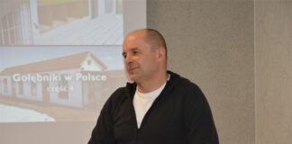 Obraz zawiera: Krótko obcięty mężczyzna w średnim wieku ubrany w ciemną bluzę polarową stoi na tle ekranu projekcyjnego, na którym pokazane są gołębniki w Polsce.