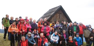 Grupa turystów wspólnie pozuje do zdjęcia. W tle widać zieloną polanę i drewnianą kapliczkę.