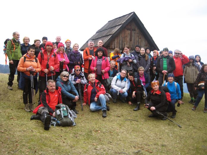 Grupa turystów wspólnie pozuje do zdjęcia. W tle widać zieloną polanę i drewnianą kapliczkę.