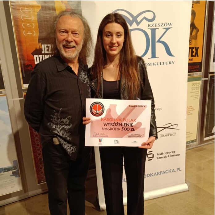 na zdjęciu stoi starszy mężczyzna i młoda dziewczyna, solistka jasielskiego domu kultury. on ma dłuższe siwe włosy i siwą brodę, ona ciemne długie włosy. Obydwoje ubrani są na czarno, dziewczyna trzyma przed sobą dyplom z wyróżnieniem w konkursie. Za nimi rollap wojewódzkiego domu kultury w rzeszowie, niebieskie napisy na białym tle. Obydwoje się uśmiechają