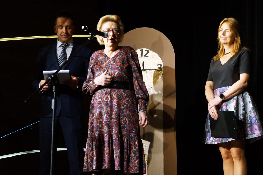 Na scenie stoi pani Krystyna Dzik, dyrektor Ośrodka Szkolno-Wychowawczego w Jaśle i wita zgromadzoną publiczność. Pani dyrektor towarzyszy para prowadzących: kobieta i mężczyzna.