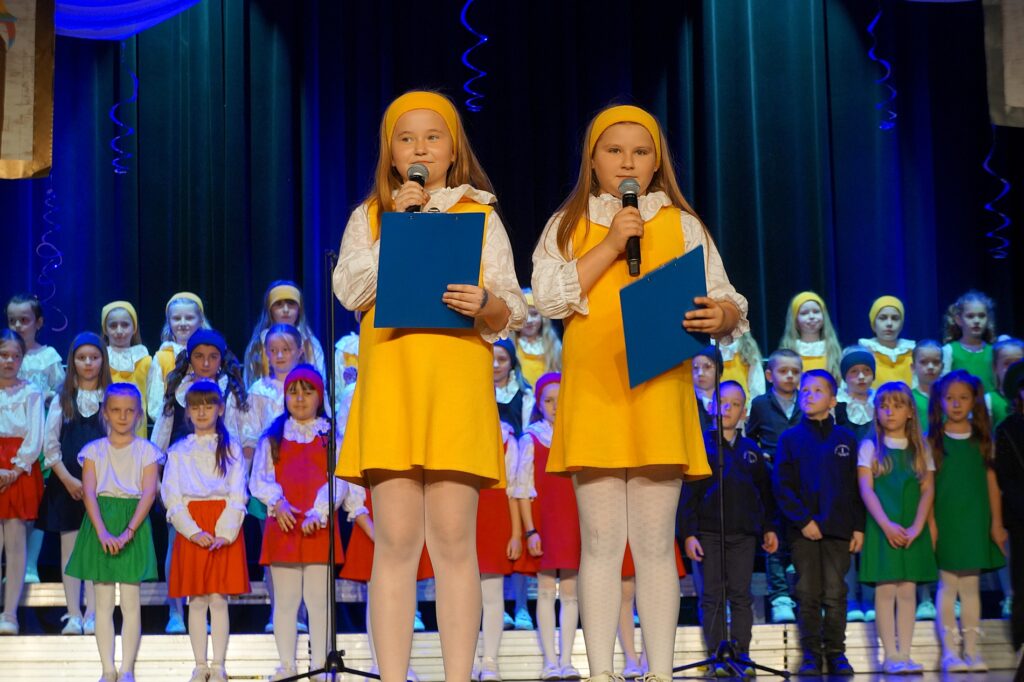 Dwie dziewczynki ubrane w stroje koloru żółtego stoją na scenie wspólnie zapowiadają występ zespołu ,,Świetliki''.