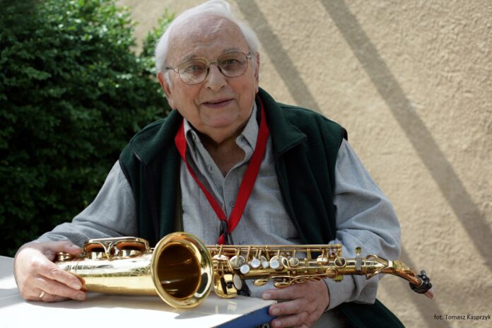 Zdjęcie: starszy mężczyzna Jerzy Duduś Matuszkiewicz siedzi na zewnątrz, przed nim na stole leży saksofon. Mężczyzna delikatnie się uśmiecha