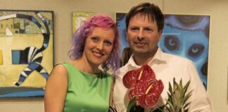 Galeria jasielskiego domu kultury. Dwoje ludzi w średnim wieku, mężczyzna i kobieta. Za nimi w tle obrazy. Obydwoje pozują do zdjęcia uśmiechnięci i trzymają kwiaty w rękach