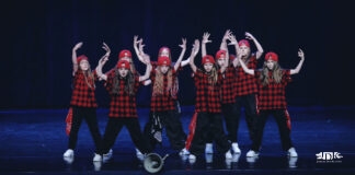 Tancerki JDK na podium Na ciemnoniebieskiej scenie pozują do zdjęcia dziewczyny z grupy tanecznej jasielskiego domu kultury. Na głowie mają czerwone czapki odwrócone daszkiem do tyłu, ubrane są w koszule w czarnoczerwoną kratę, czarne spodnie i białe buty. trzymają ręce wzniesione do góry. Przed nimi na scenie leży głośnik