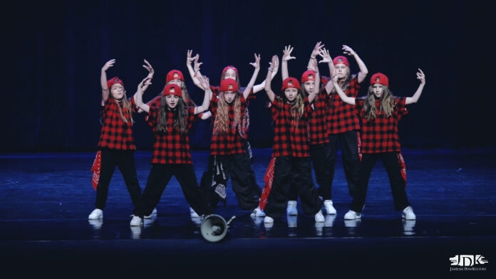 RR Kids Intermedium Na ciemnoniebieskiej scenie pozują do zdjęcia dziewczyny z grupy tanecznej jasielskiego domu kultury. Na głowie mają czerwone czapki odwrócone daszkiem do tyłu, ubrane są w koszule w czarnoczerwoną kratę, czarne spodnie i białe buty. trzymają ręce wzniesione do góry. Przed nimi na scenie leży głośnik