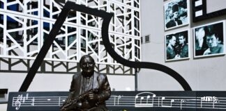 Skwerek jasielskiego domu kultury, a na nim muzyczna ławeczka jerzego dudusia matuszkiewicza. postać z brązu w naturalnych rozmiarach siedzi na ławce i trzyma w dłoniach saksofon. Obok leżą prawdziwie słoneczniki