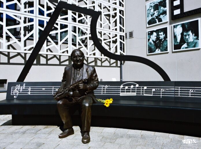 Muzyczna ławeczka Jerzego Matuszkiewicza Skwerek jasielskiego domu kultury, a na nim muzyczna ławeczka jerzego dudusia matuszkiewicza. postać z brązu w naturalnych rozmiarach siedzi na ławce i trzyma w dłoniach saksofon. Obok leżą prawdziwie słoneczniki