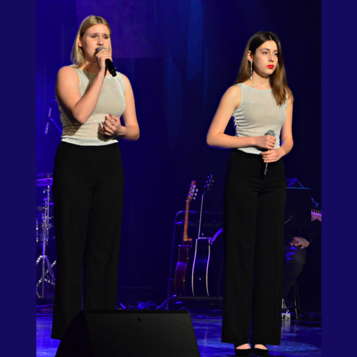 Laura Gondek i Karolina Polak Na scenie stoją dwie dziewczyny, mają jasne bluzki bez rękawów i czarne długie spodnie. Śpiewają do mikrofonów, scena jest w ciemnej scenerii