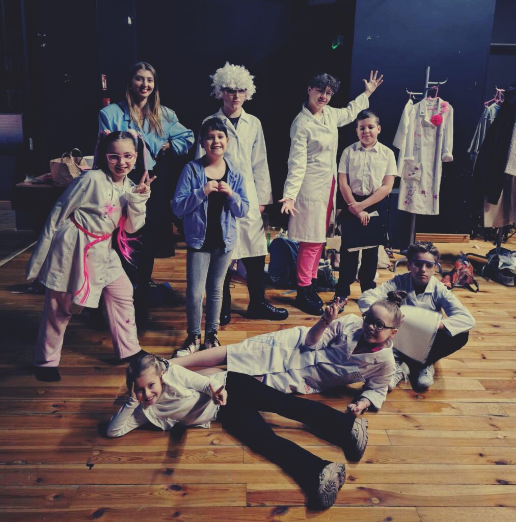 dziecięca grupa teatralna za kulisami sceny. część z nich stoi, a część leży na drewnianej podłodze