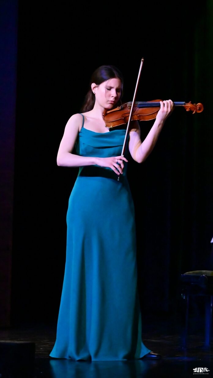 DSC_6585 Młoda kobieta w długiej, eleganckiej, zielonej sukni gra na skrzypcach na scenie. Jej twarz wyraża głębokie skupienie, a postawa jest pełna gracji. Ciemne tło sceny podkreśla jej sylwetkę i instrument, nadając występowi wyjątkowy nastrój.
