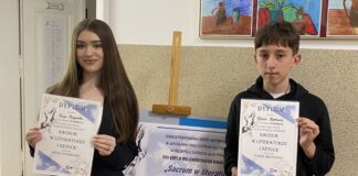 25 Wojewódzki Konkurs „Sacrum w Literaturze i sztuce” – główne nagrody dla wokalistów JDK Dwoje młodych ludzi stoi w korytarzu szkolnym, trzymając dyplomy. Dziewczyna po lewej stronie ma długie, ciemne włosy i jest ubrana w elegancki, czarny garnitur. Chłopak po prawej stronie nosi czarną bluzę i spodnie dresowe z białymi napisami. Oboje uśmiechają się delikatnie i prezentują swoje wyróżnienia za udział w konkursie „Sacrum w literaturze i sztuce” w kategorii poezji śpiewanej. W tle znajduje się tablica informacyjna z plakatem konkursowym oraz kolorowe prace plastyczne zawieszone na ścianie.