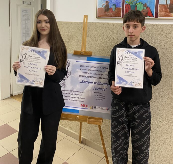 Dwoje młodych ludzi stoi w korytarzu szkolnym, trzymając dyplomy. Dziewczyna po lewej stronie ma długie, ciemne włosy i jest ubrana w elegancki, czarny garnitur. Chłopak po prawej stronie nosi czarną bluzę i spodnie dresowe z białymi napisami. Oboje uśmiechają się delikatnie i prezentują swoje wyróżnienia za udział w konkursie „Sacrum w literaturze i sztuce” w kategorii poezji śpiewanej. W tle znajduje się tablica informacyjna z plakatem konkursowym oraz kolorowe prace plastyczne zawieszone na ścianie.