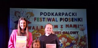 Nagrody i wyróżnienia dla wokalistek JDK w Przemyślu Trzy dziewczyny stoją na scenie podczas Podkarpackiego Festiwalu Piosenki „Śpiewaj Razem z Nami”. Każda z nich trzyma dyplom z wyróżnieniem. Najstarsza, po lewej, ubrana jest w różowy garnitur i trzyma dyplom z napisem „I NAGRODA”. Najmłodsza, w białej sukience i czarnych balerinach, trzyma dyplom „III NAGRODA”. Trzecia dziewczyna, w czarnej bluzce i zielonej spódnicy, ma dyplom z wyróżnieniem. W tle znajduje się duży baner festiwalu z kolorową grafiką i napisami, a na scenie ustawione są instrumenty muzyczne, w tym perkusja.