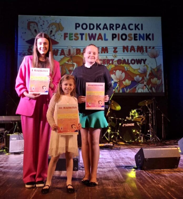 Finał Śpiewaj razem z nami Trzy dziewczyny stoją na scenie podczas Podkarpackiego Festiwalu Piosenki „Śpiewaj Razem z Nami”. Każda z nich trzyma dyplom z wyróżnieniem. Najstarsza, po lewej, ubrana jest w różowy garnitur i trzyma dyplom z napisem „I NAGRODA”. Najmłodsza, w białej sukience i czarnych balerinach, trzyma dyplom „III NAGRODA”. Trzecia dziewczyna, w czarnej bluzce i zielonej spódnicy, ma dyplom z wyróżnieniem. W tle znajduje się duży baner festiwalu z kolorową grafiką i napisami, a na scenie ustawione są instrumenty muzyczne, w tym perkusja.