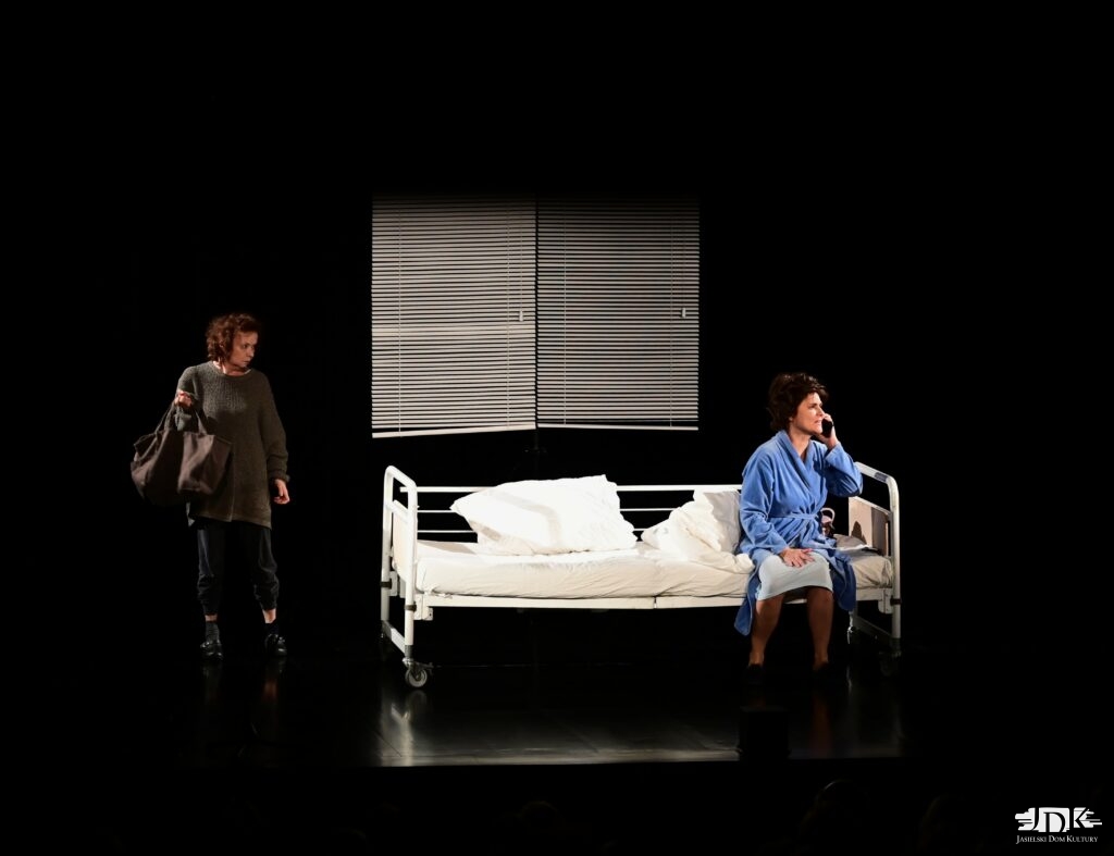 jedna kobieta siedząca na łóżku, druga stojąca po lewej stronie