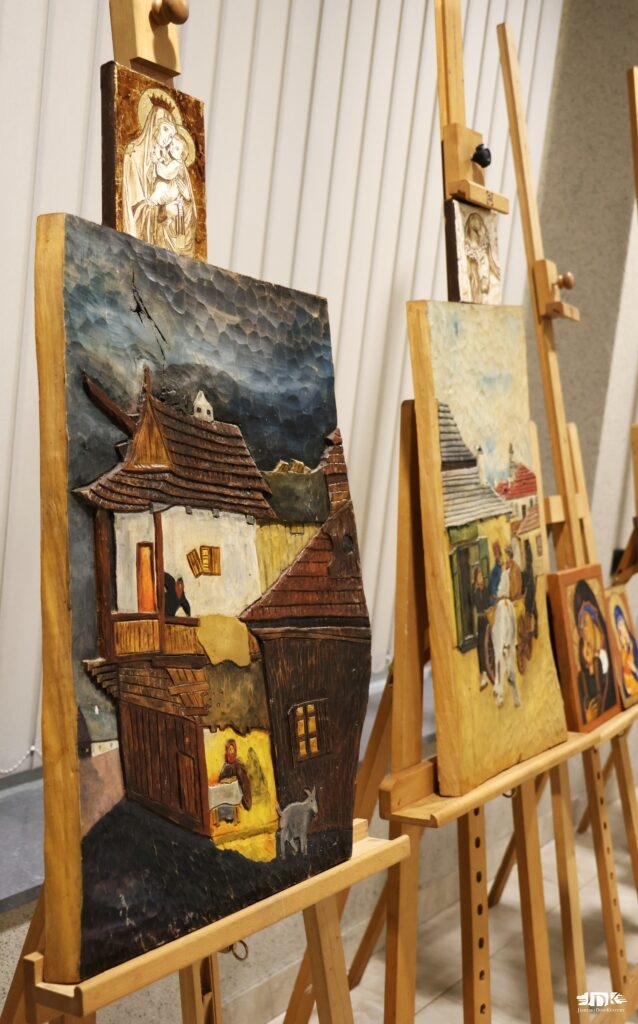 rzeźba w drewnie malowana w kolorze, przedstawiająca scenkę: wysoki drewniano-murowany dom, na dole domu warsztat lub izba, na górze na balkonie stoi mężczyzna