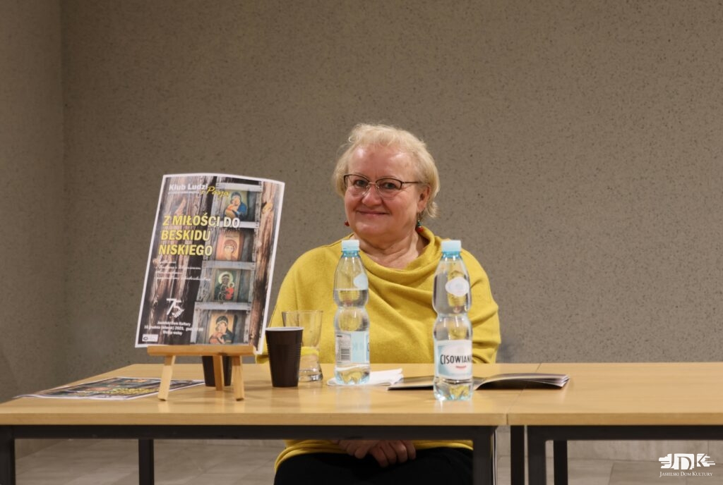 uśmiechnięta Pani Marchewka w żółtym swetrze siedzi przy stoliku. Po lewej stornie zdjęcia widoczny plakat z wydarzenia