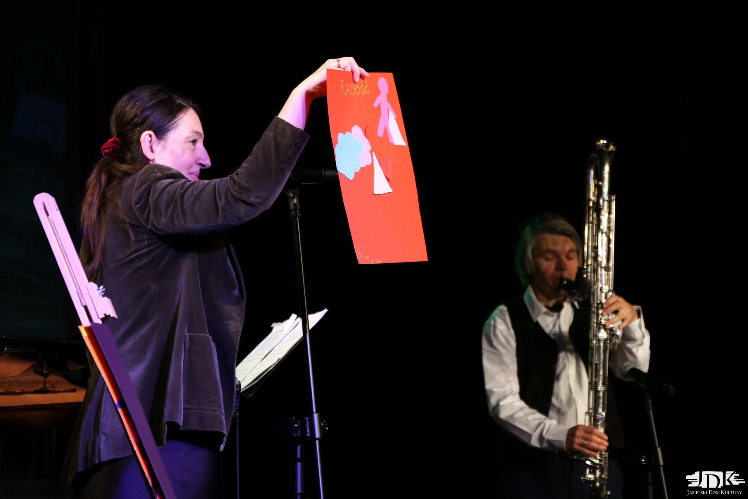 skrzypaczka trzymająca czerwony arkusz z wycinankami, a w tle klarnecista