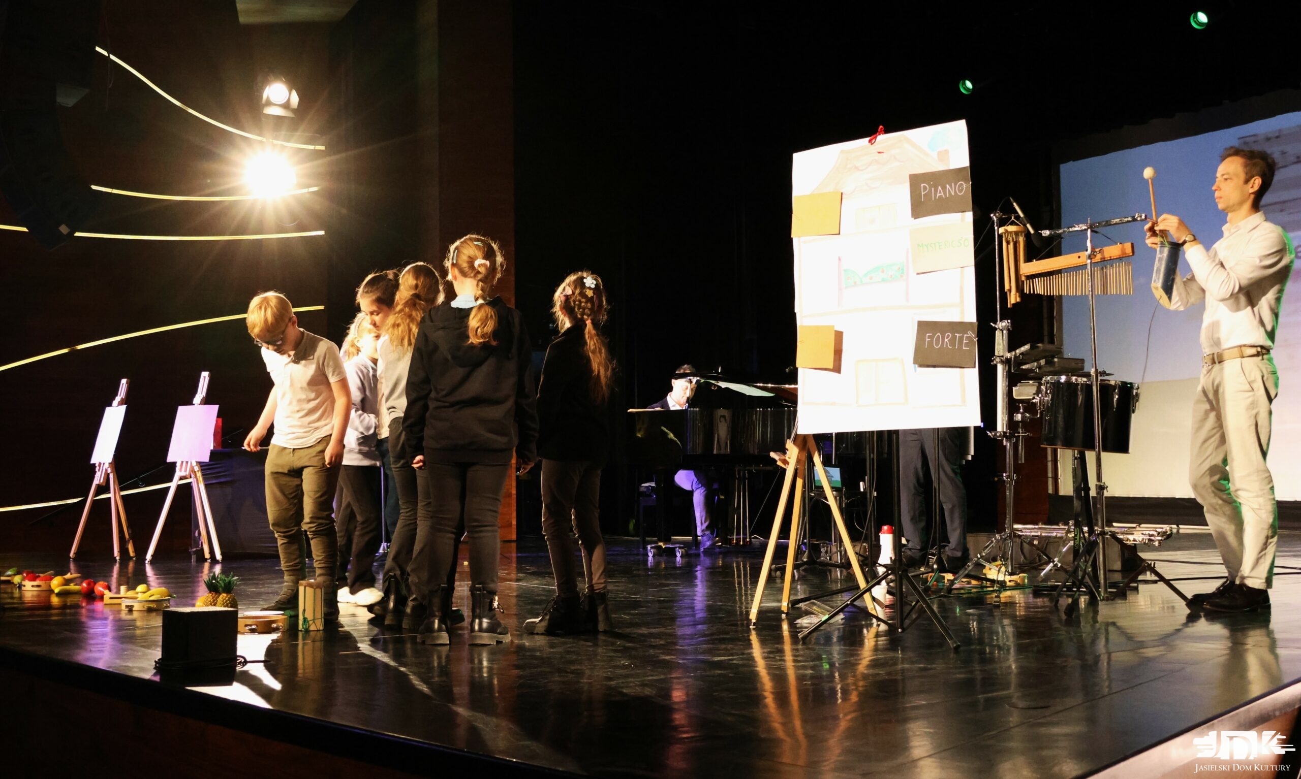 na scenie grupka dzieci a w tle instrumenty perkusyjne, fortepian i arkusz papieru z namalowanym domem