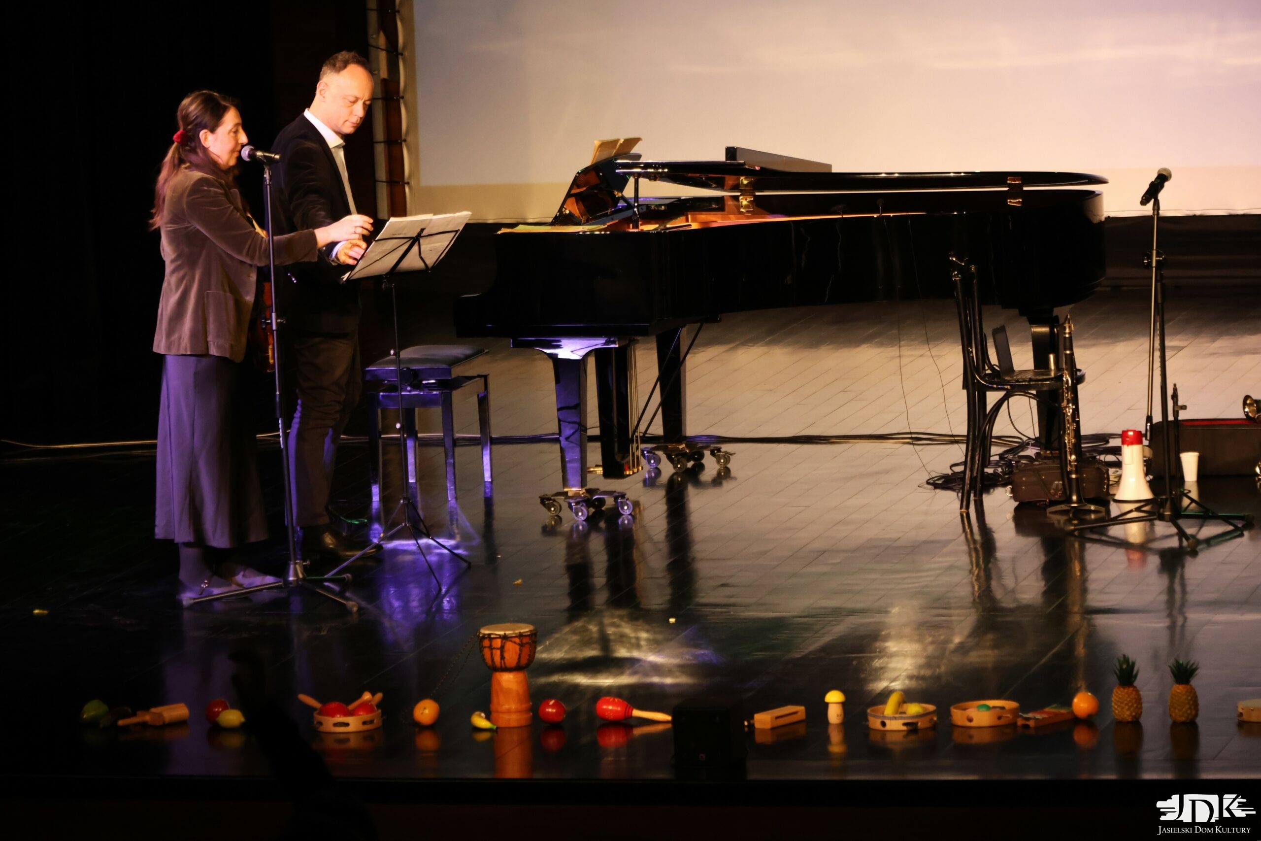 skrzypaczka mówiąca do mikrofonu oraz muzyk przy fortepianie. Na dole sceny instrumenty: grzechotki, tamburyna, bongosy