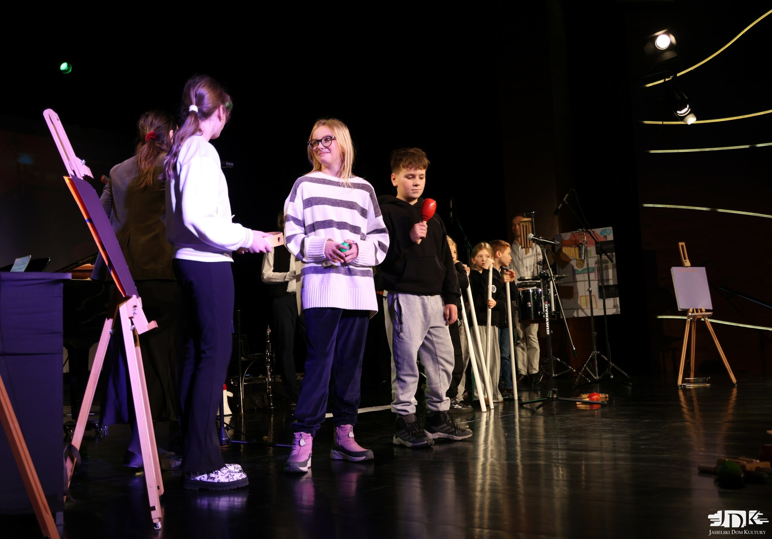 grupka dzieci na scenie ustawiona w linii, trzymająca w rękach grzechotki, tuby i tamburyn