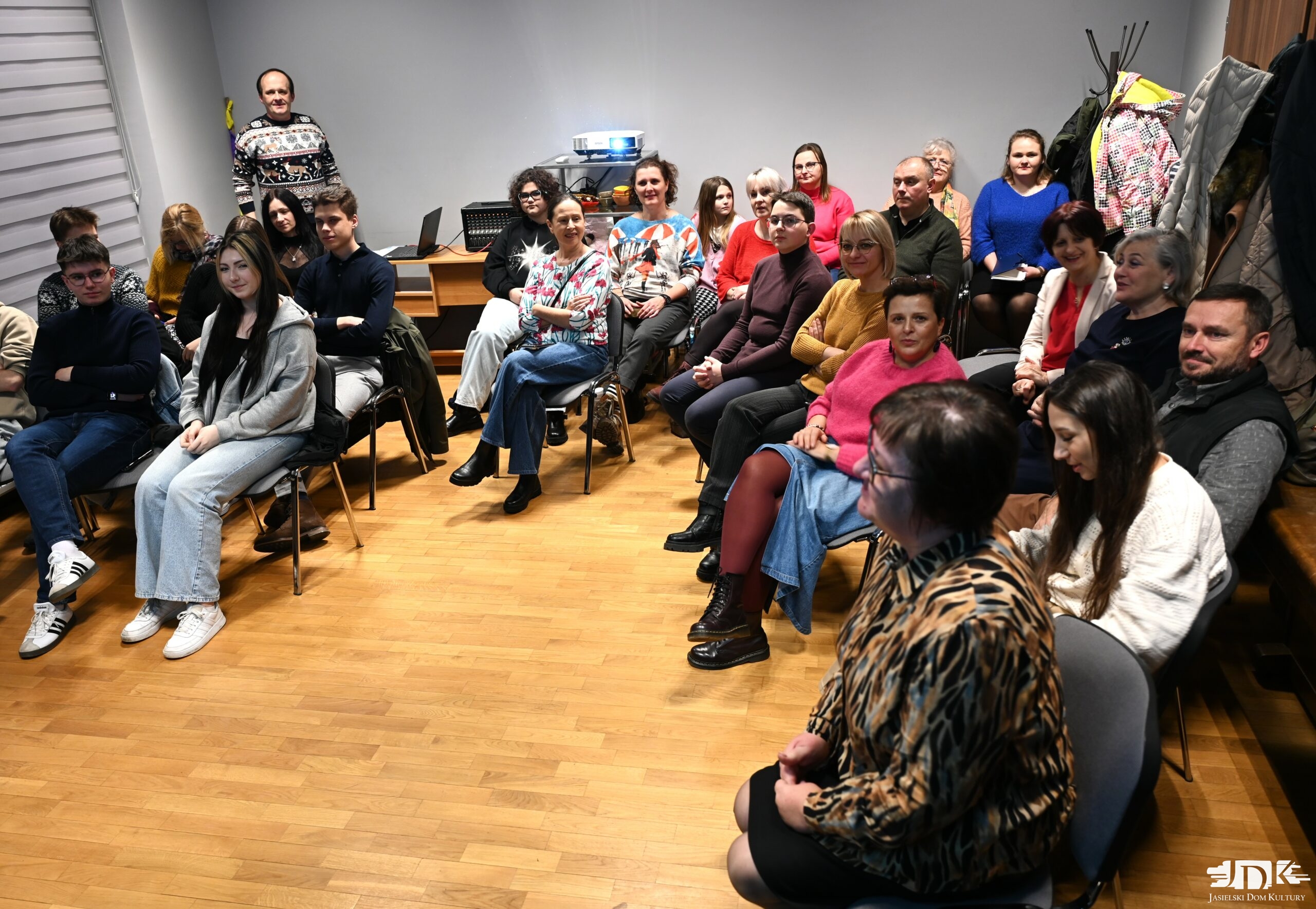 grupa uczestników w różnym wieku siedząca wokół sali, patrząca w aparat