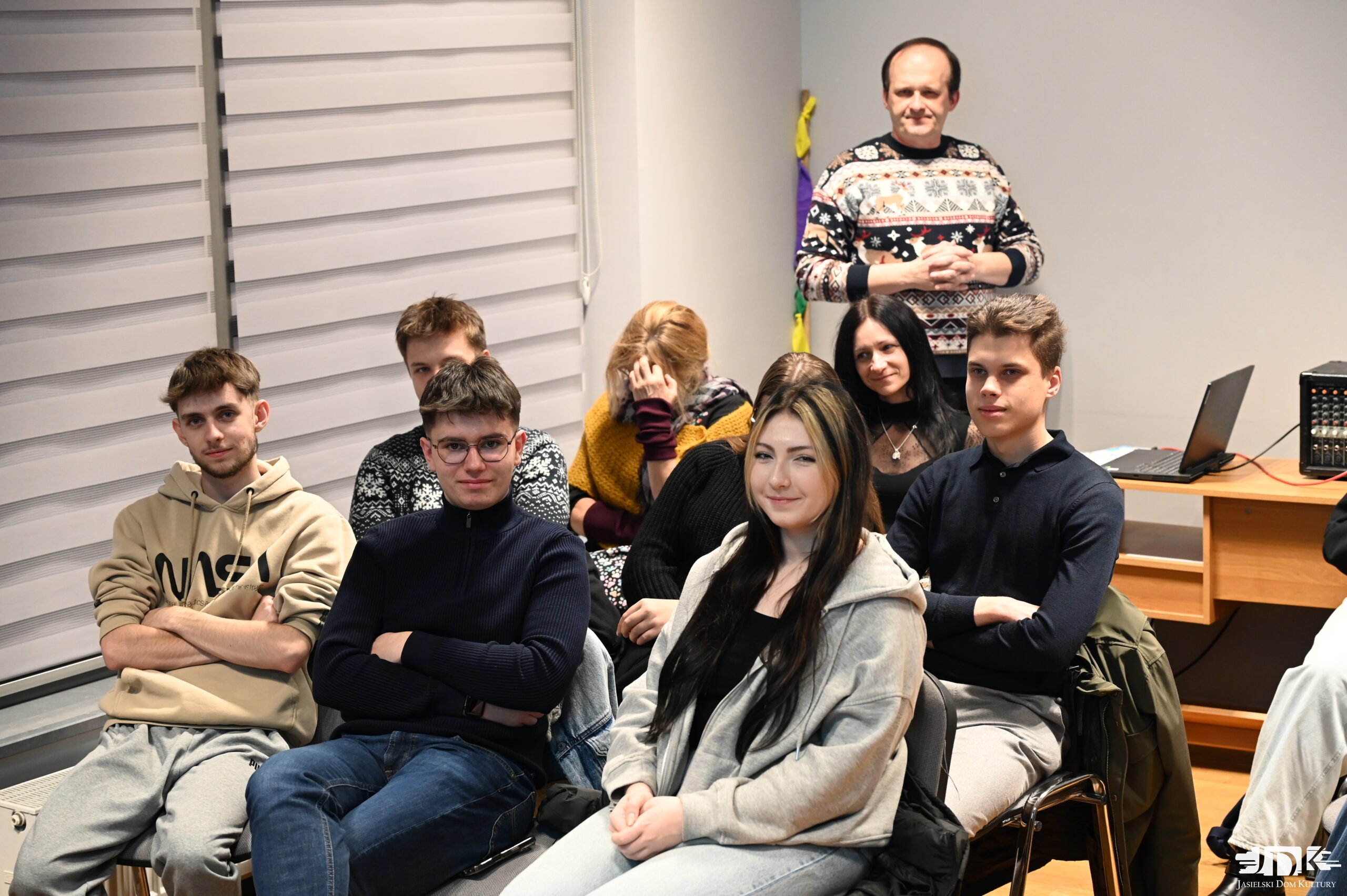 troje młodych ludzi w bluzach dresowych: dwóch chłopaków i jedna dziewczyna patrzący w obiektyw. Za nimi pięcioro innych uczestników, a w tle Bartosz Gałązka