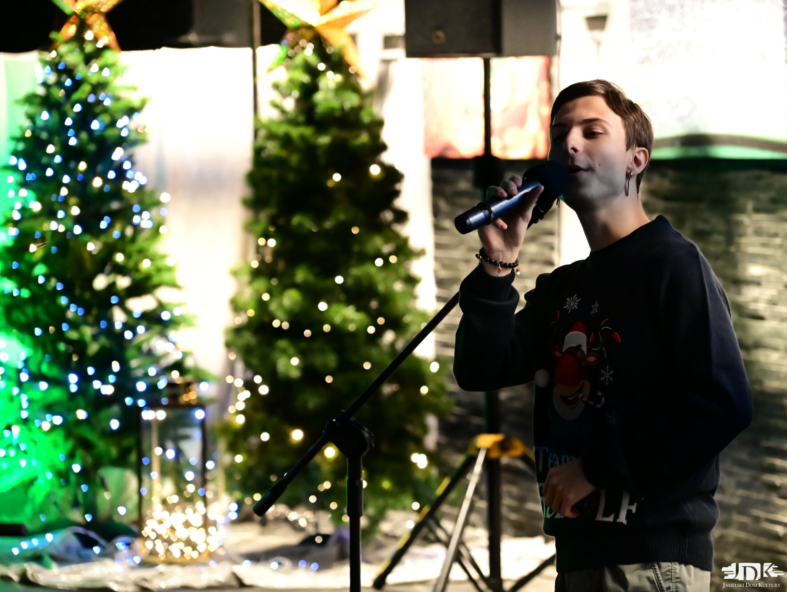 prowadzący domówkę w swetrze z reniferem z czerwonym nosem, śpiewający do mikrofonu. W tle dwie choinki