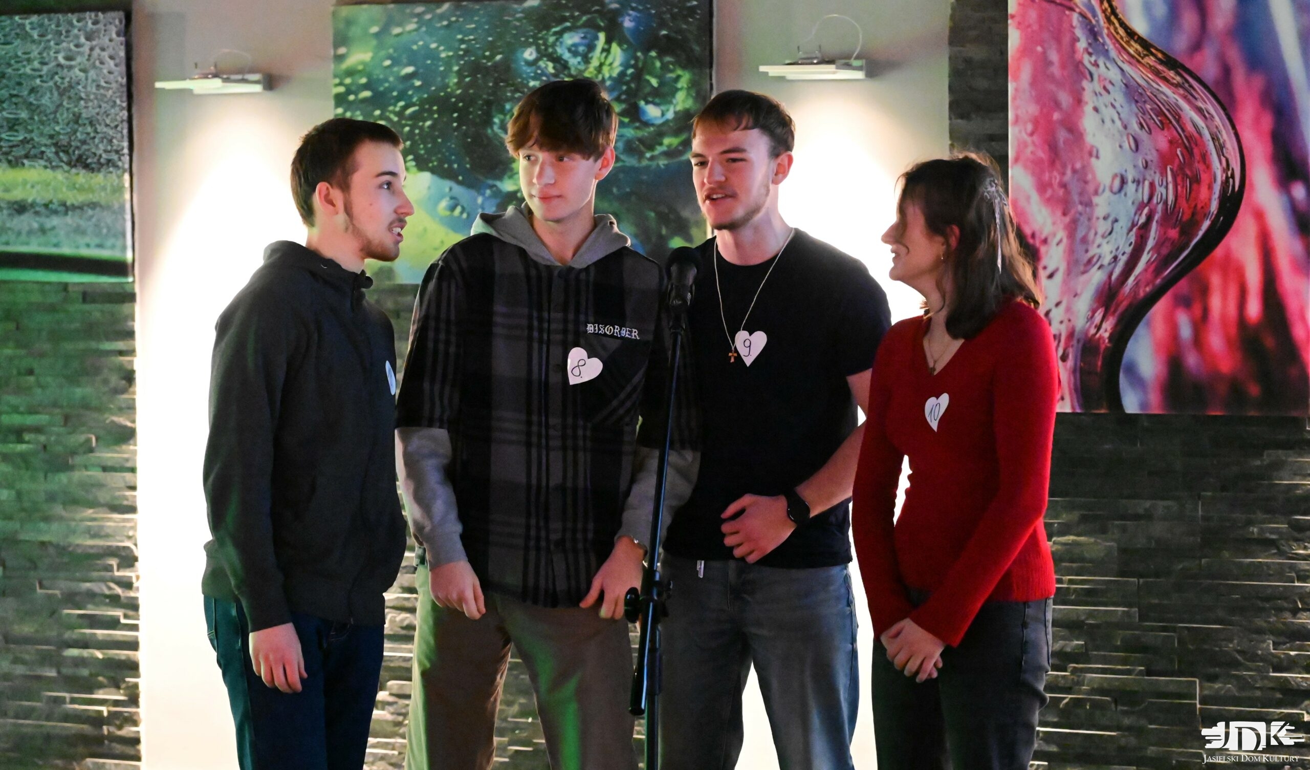 czwórka znajomych śpiewa do mikrofonu: 3 chłopaków ubranych na czarno i dziewczyna w czerwonej bluzce