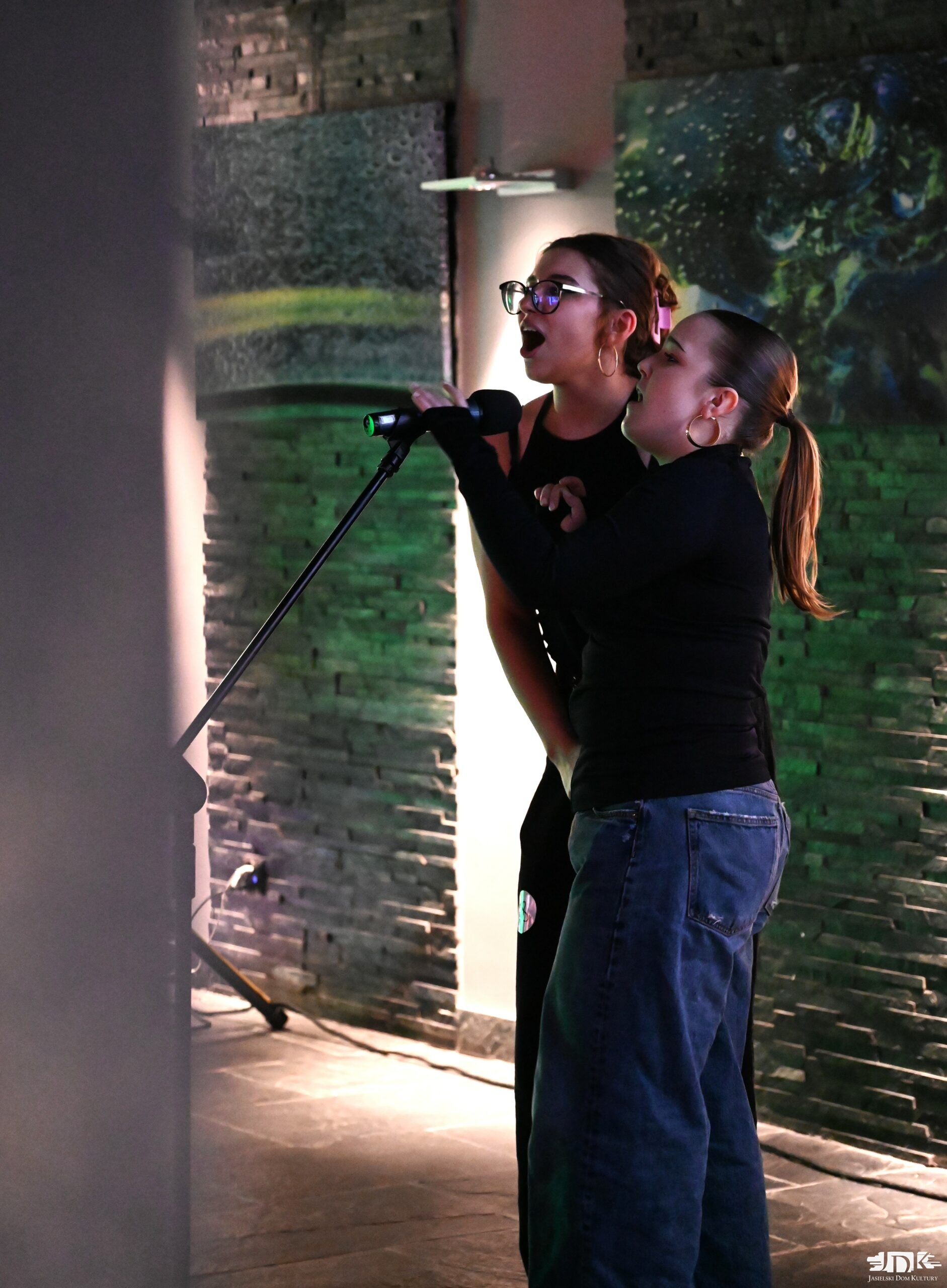 dwie dziewczyny: jedna w kucyku w dżinsach i czarnej bluzce oraz druga w okularach cała ubrana na czarno śpiewają do mikrofonu