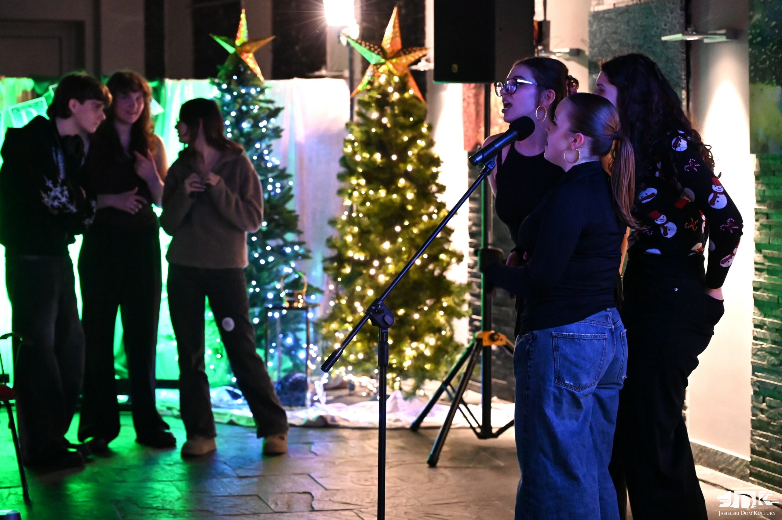 trzy uczestniczki śpiewają do mikrofonu, w tle troje znajomych stoi i rozmawia tuz przy choinkach
