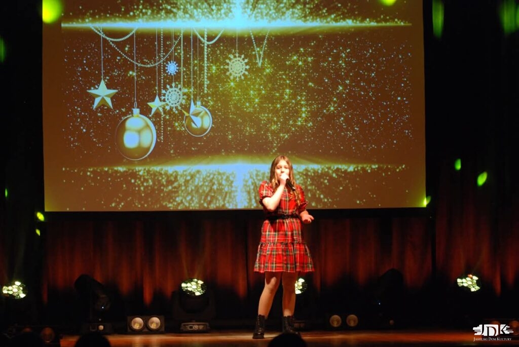 dzieewczynka w czerwonej sukience na scenie śpiewająca. w tle czarno-złoty ekran z bombkami