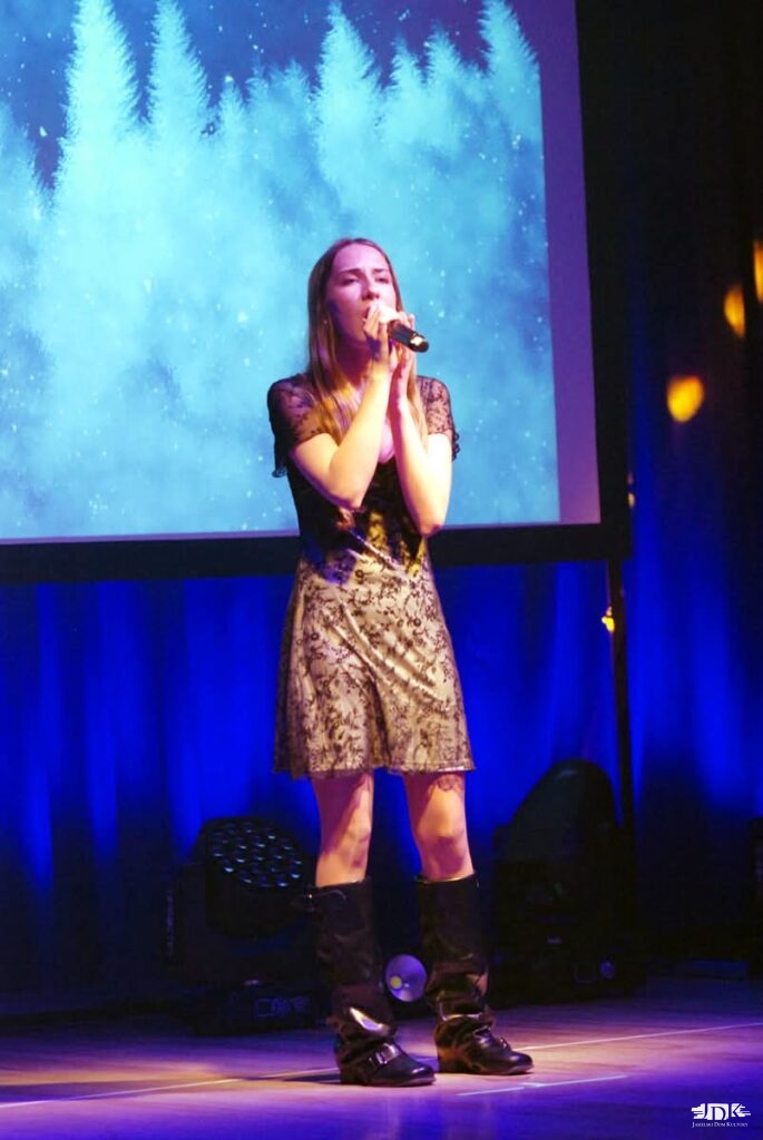 dziewczynka w złotej sukience śpiewa, w tle niebieski materiał i szary ekran