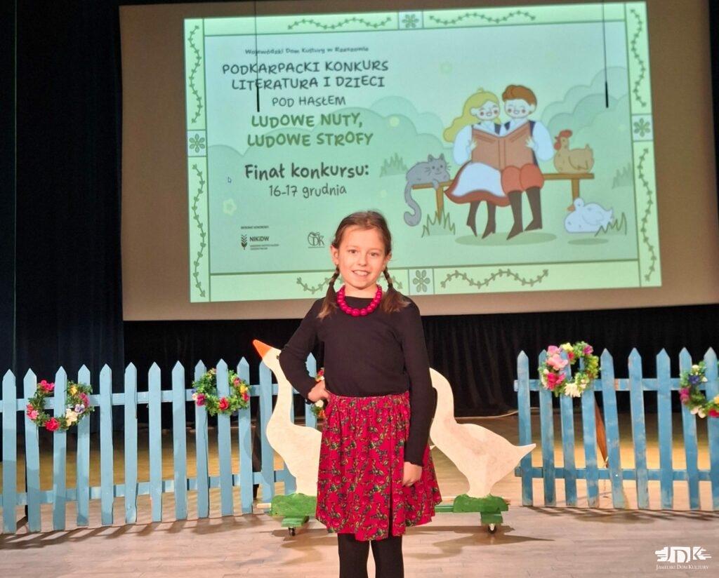 wokalistka w stroju: czerwona spódnica w kwiaty oraz czarna bluzka z czerwonymi koralami. Za nią niebieski płotek oraz makiety gęsi