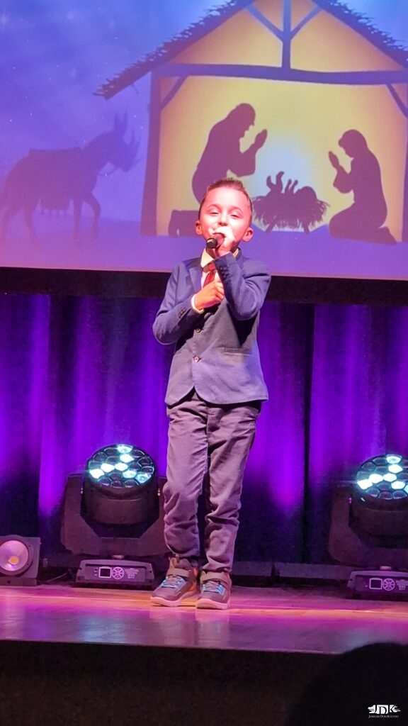 chłopiec w szarych spodniach i szarej marynarce śpiewa, spoglądając na widownię