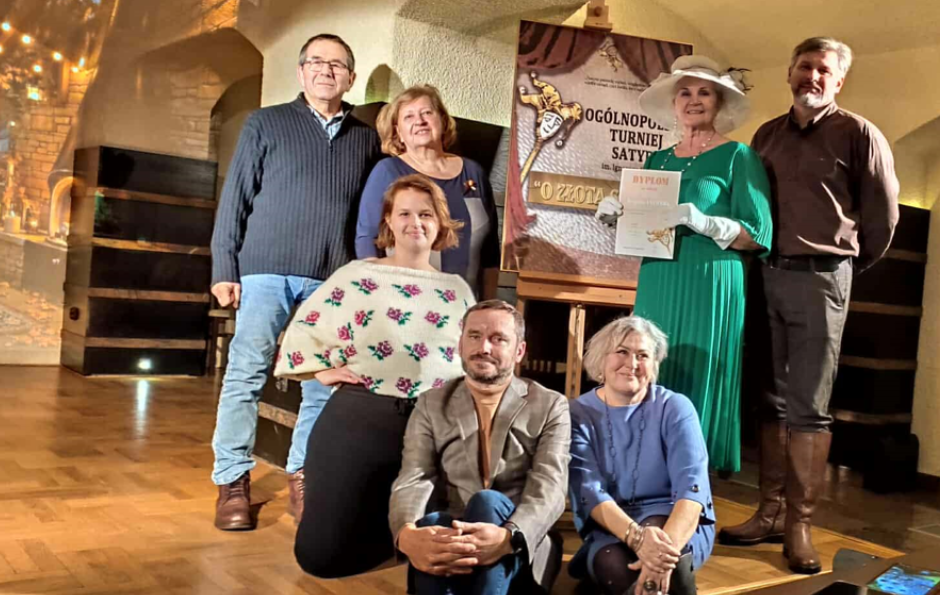 członkowie grupy teatralnej w kolorowych ubraniach pozują z dyplomem oraz plakatem konkursu