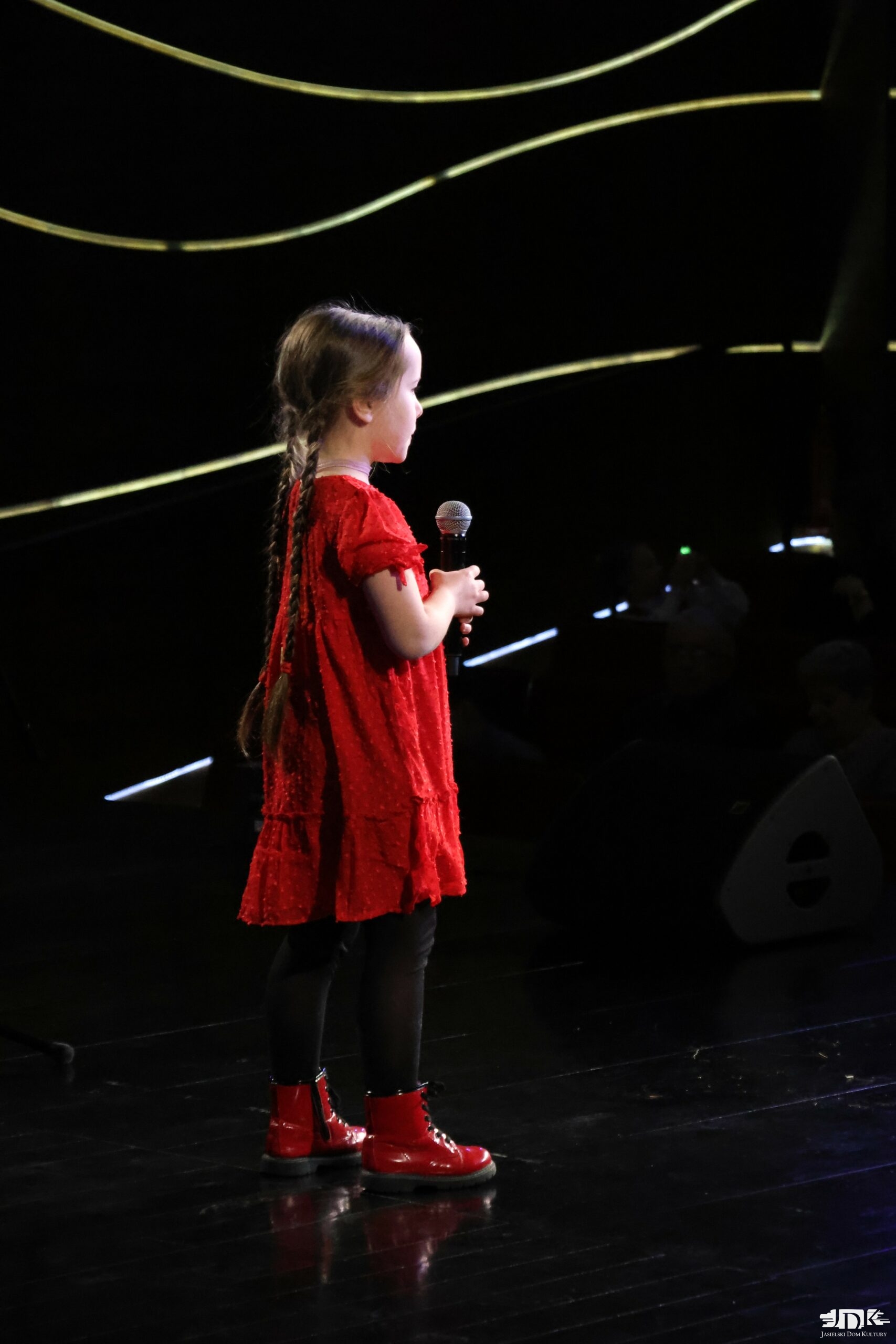 dziewczynka z mikrofonem w dłoni, stojąca bokiem do kamery. W warkoczach i czerwonej sukience