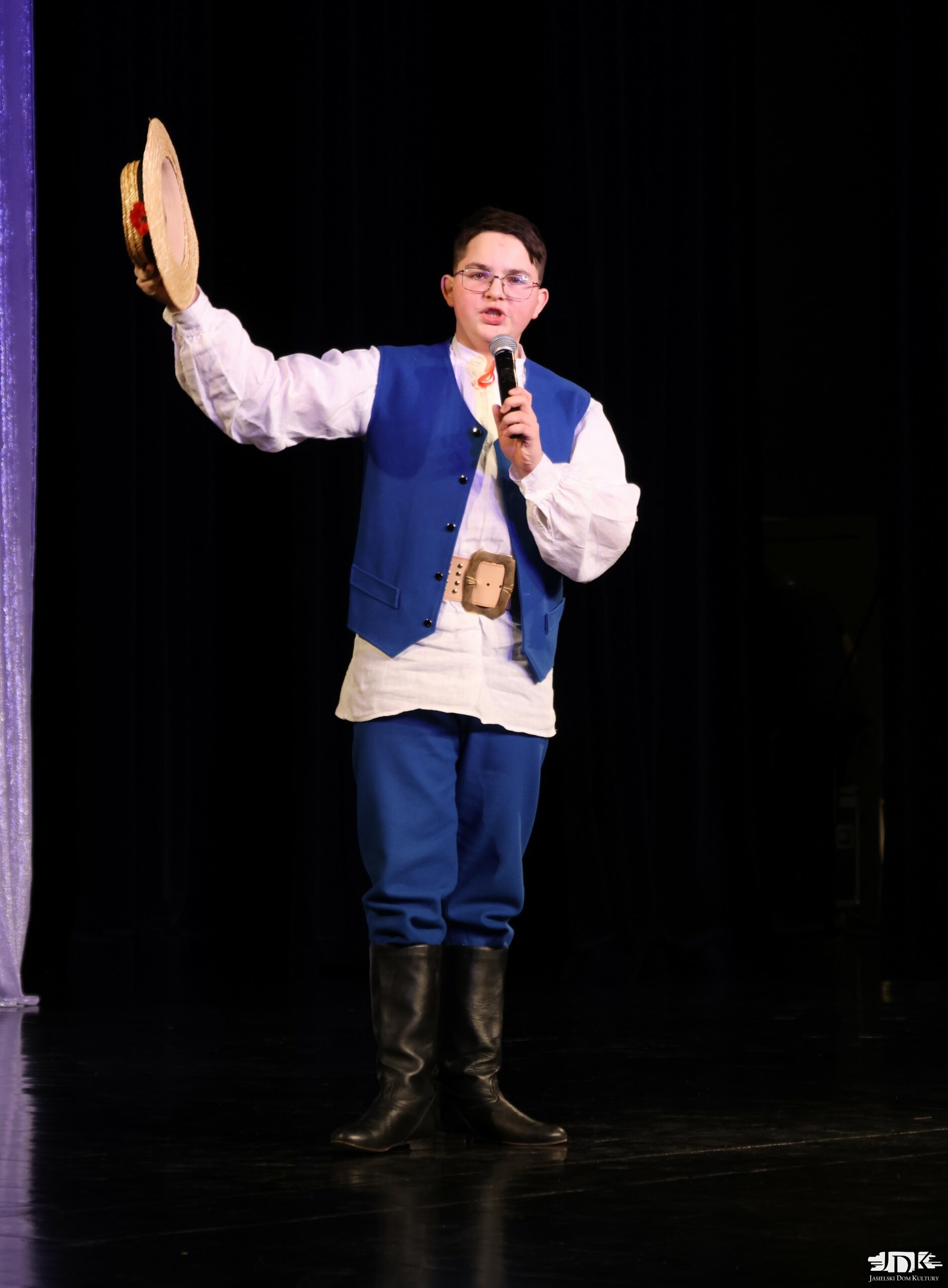 chłopiec w ludowym stroju trzymający w uniesionej ręce słomkowy kapelusz