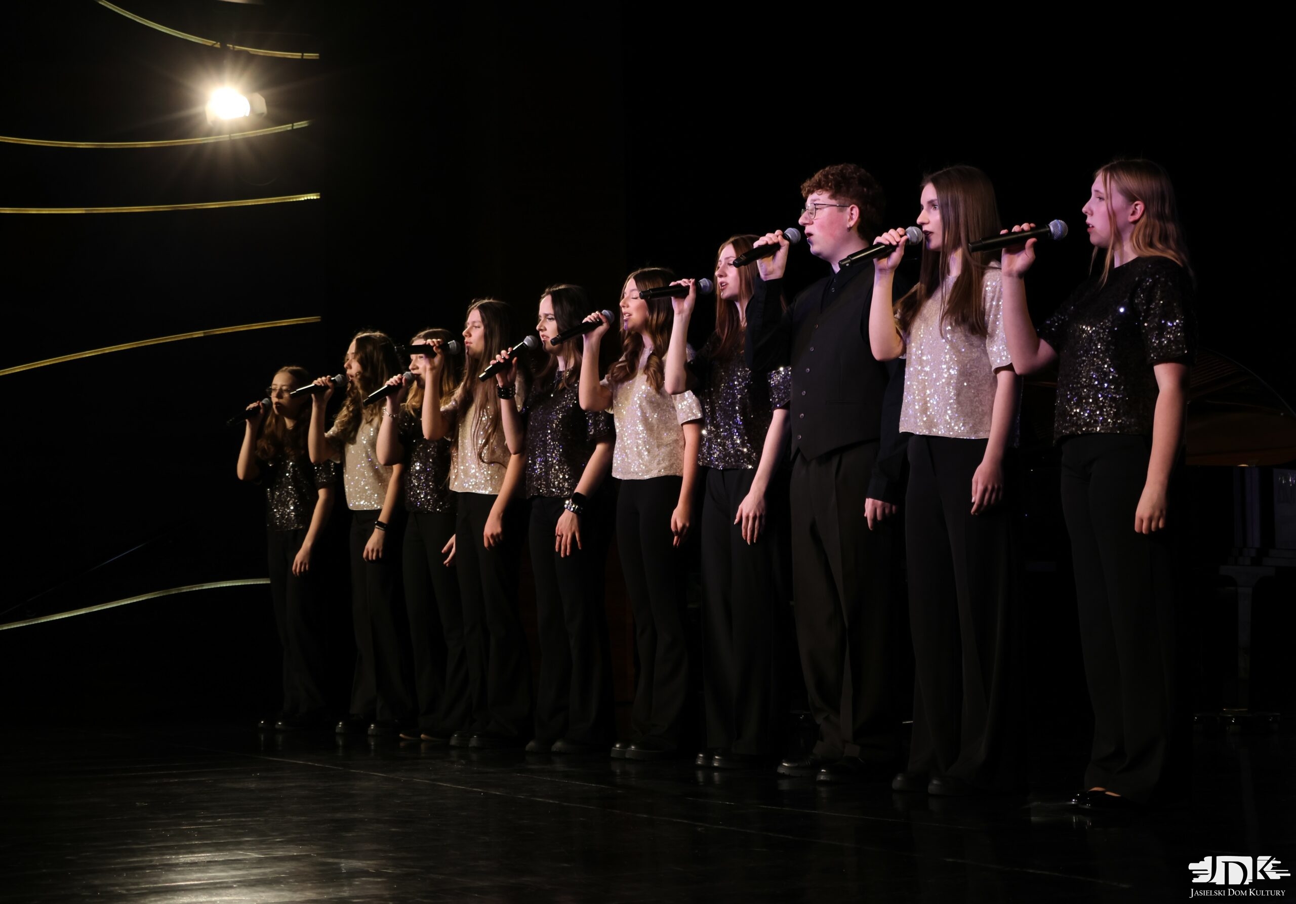 zespół wokalny w białych i czarnych cekinowych bluzkach, ustawiony naprzemiennie. 9 dziewcząt i jeden chłopiec